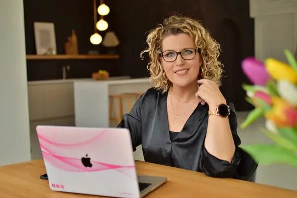 Een vrouw met krullend blond haar en een bril zit aan een tafel met een laptop en lacht naar de camera. Een vaas met kleurrijke bloemen staat op de voorgrond, terwijl haar scherm het VR-ander Footer-ontwerp toont.