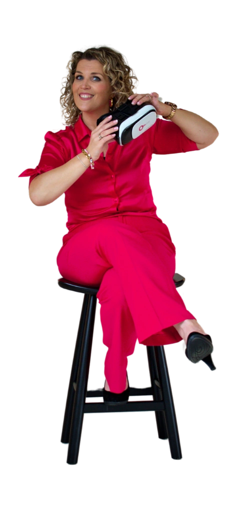 Vrouw in roze outfit zittend op een kruk, met VR-bril in haar hand, glimlachend en naar links kijkend.