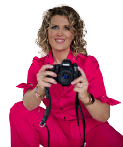 Een vrouw met krullend blond haar, gekleed in een felroze outfit, houdt een camera vast en glimlacht naar de toeschouwer.