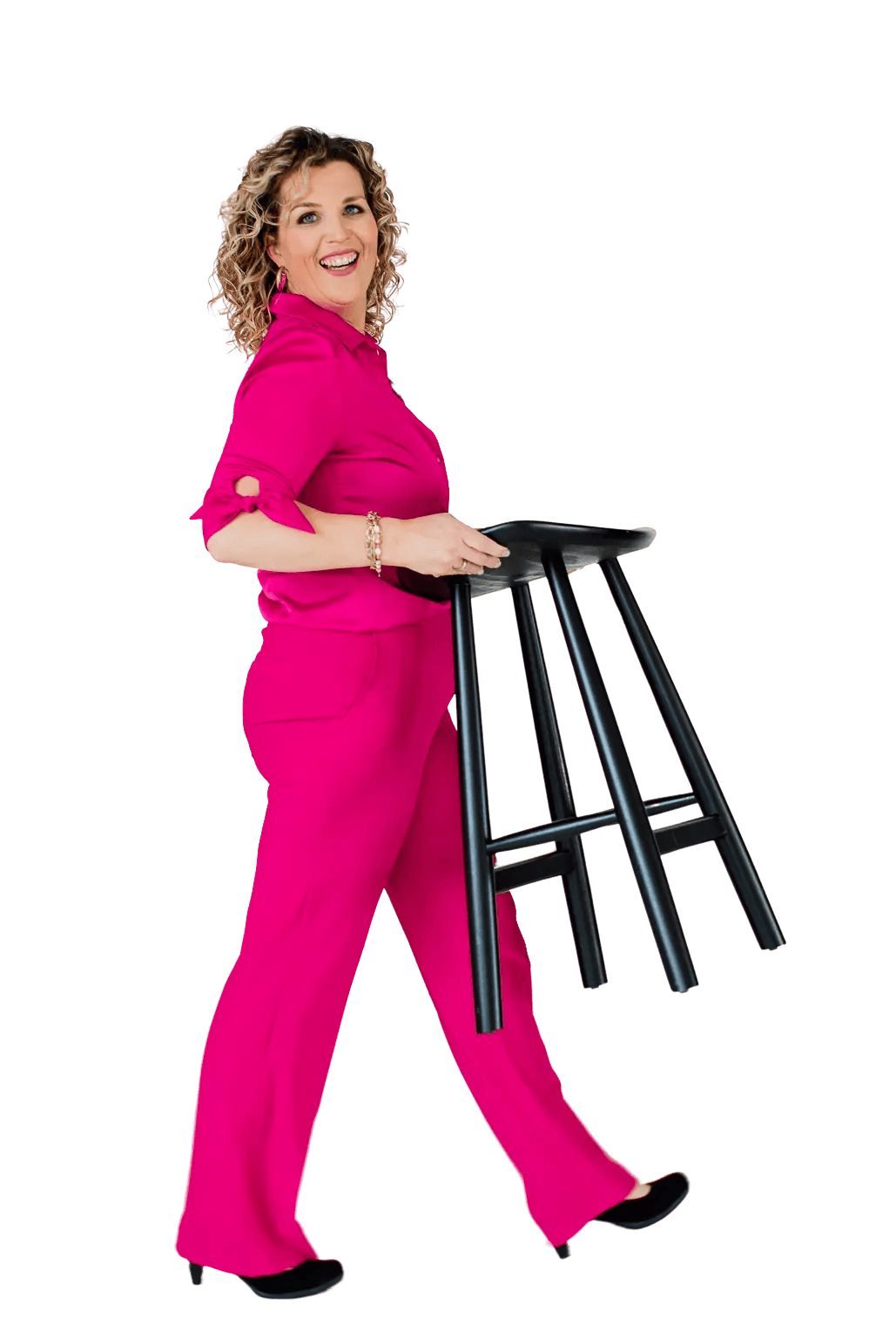 Een vrouw in een felroze broekpak staat lachend, met een zwarte kruk onder haar arm, tegen een effen achtergrond.