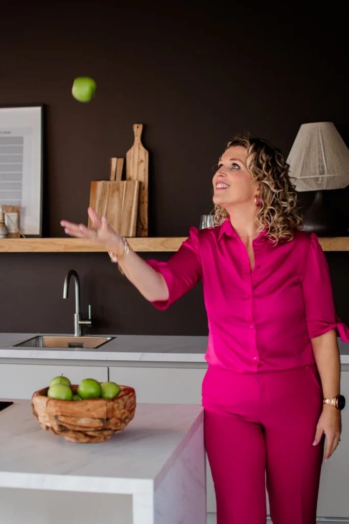 Een vrouw in een felroze outfit staat in een moderne keuken, lacht en gooit een groene appel in de lucht boven het aanrecht met een schaal vol appels.