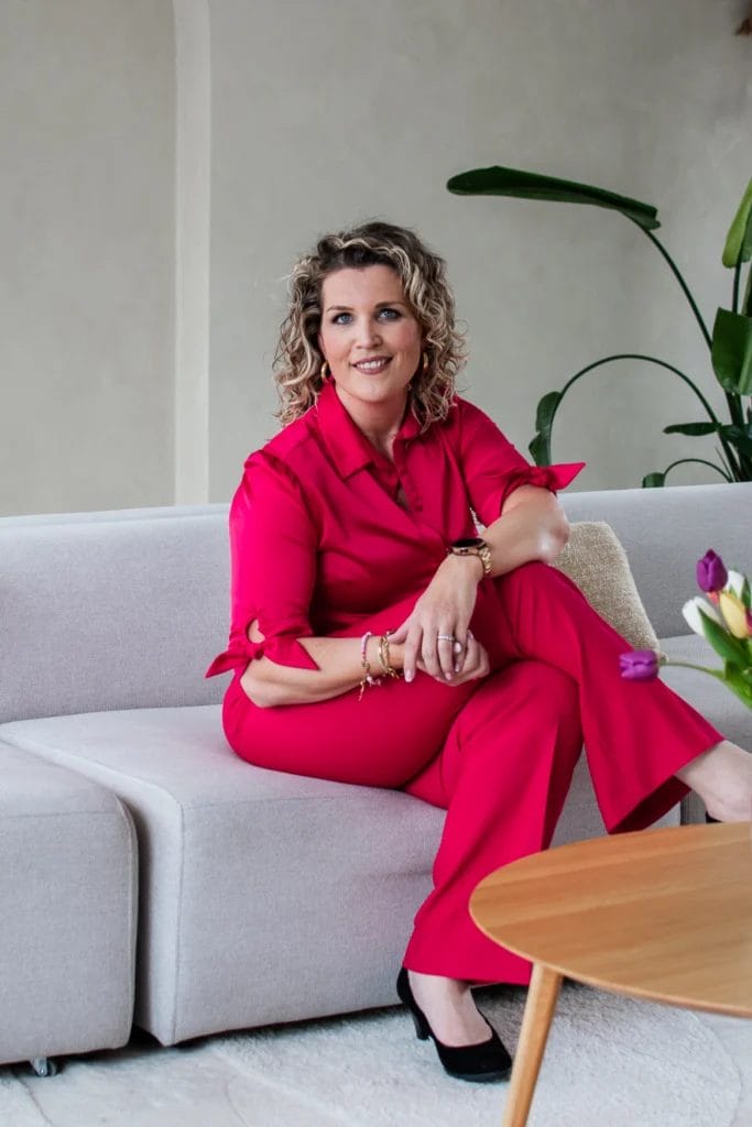 Een vrouw met krullend haar in een felroze outfit zit op een lichtgekleurde bank, warm glimlachend en met haar handen op haar knieën. Een houten tafel en planten op de achtergrond creëren een gezellige setting, perfect voor vriendelijk contact.