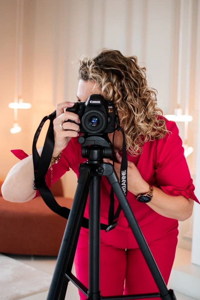 Een persoon met krullend haar in een felroze outfit maakt binnenshuis een foto voor woningfotografie met een Canon-camera op een statief.