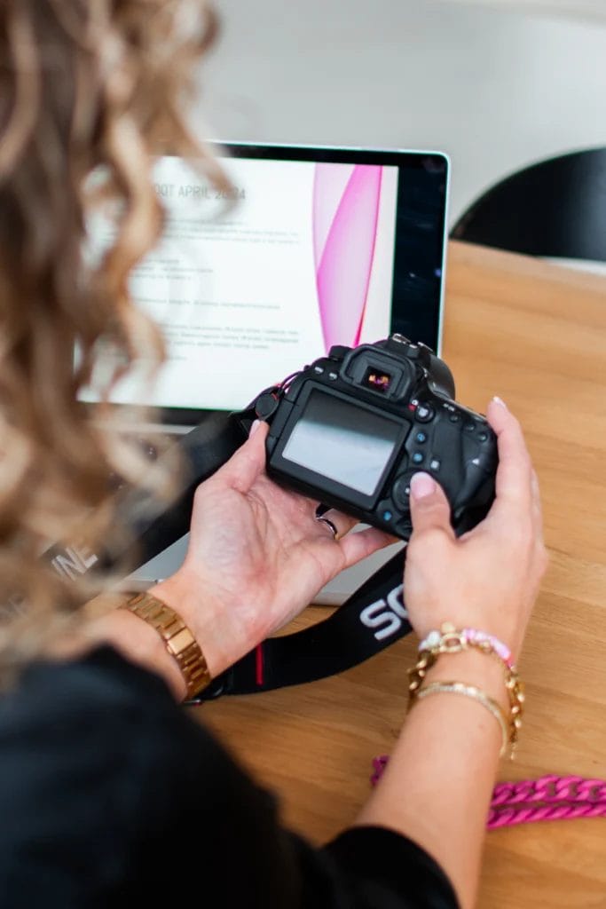 Een persoon met een digitale camera zit aan een houten tafel voor een open laptop waarop een presentatie over woningfotografie wordt getoond.
