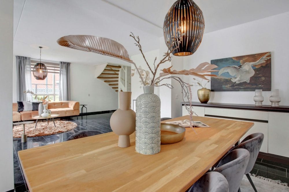 Moderne eetkamer met een houten tafel, decoratieve vazen en hanglampen. Op de achtergrond is een woonkamer met een bank en een groot schilderij van een vogel zichtbaar.