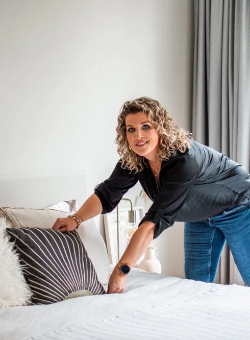 Een vrouw met krullend haar en casual kleding legt kussens op een netjes opgemaakt bed in een lichte slaapkamer met een groot raam. Hierdoor ontstaat een uitnodigende ruimte, ideaal om te ontspannen of om contact te hebben met dierbaren.