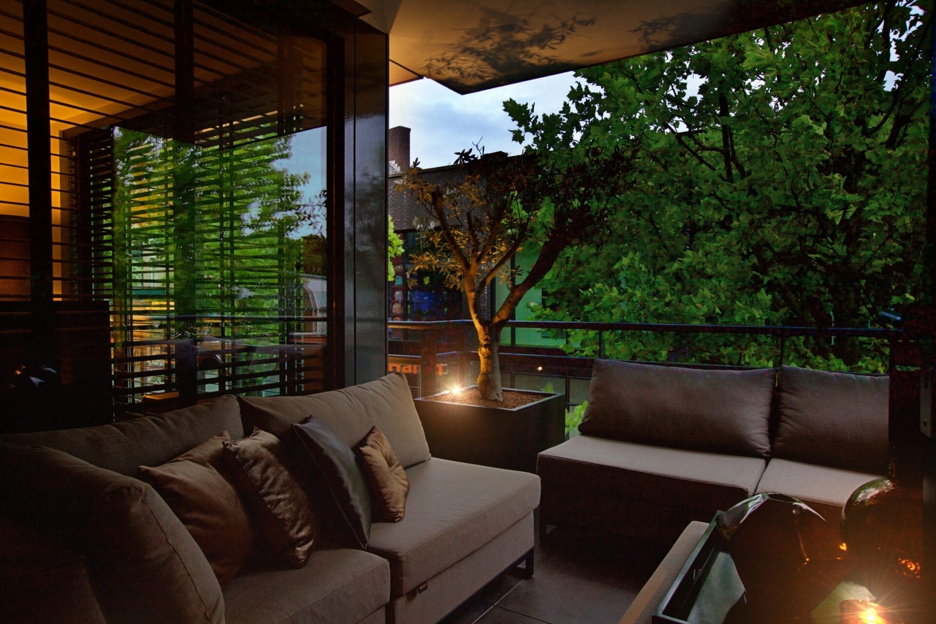 avond02 Een modern balkon met comfortabele zitplaatsen, kussens, een potplant en zachte avondverlichting, een toonbeeld van stijlvol interieurontwerp, met uitzicht op groene bomen.